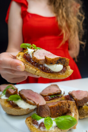 Helena is holding one duck breast sandwich with fig jam and other sandwiches on a plate
