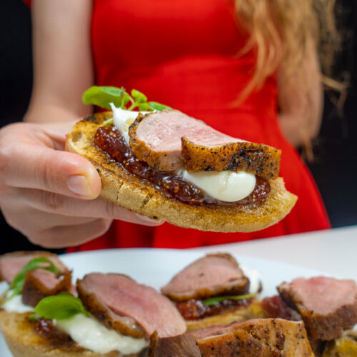 Helena is holding one duck breast sandwich with fig jam and other sandwiches on a plate