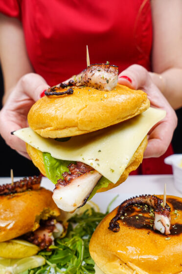 Helena is holding one open octopus burger and two burgers on a plate