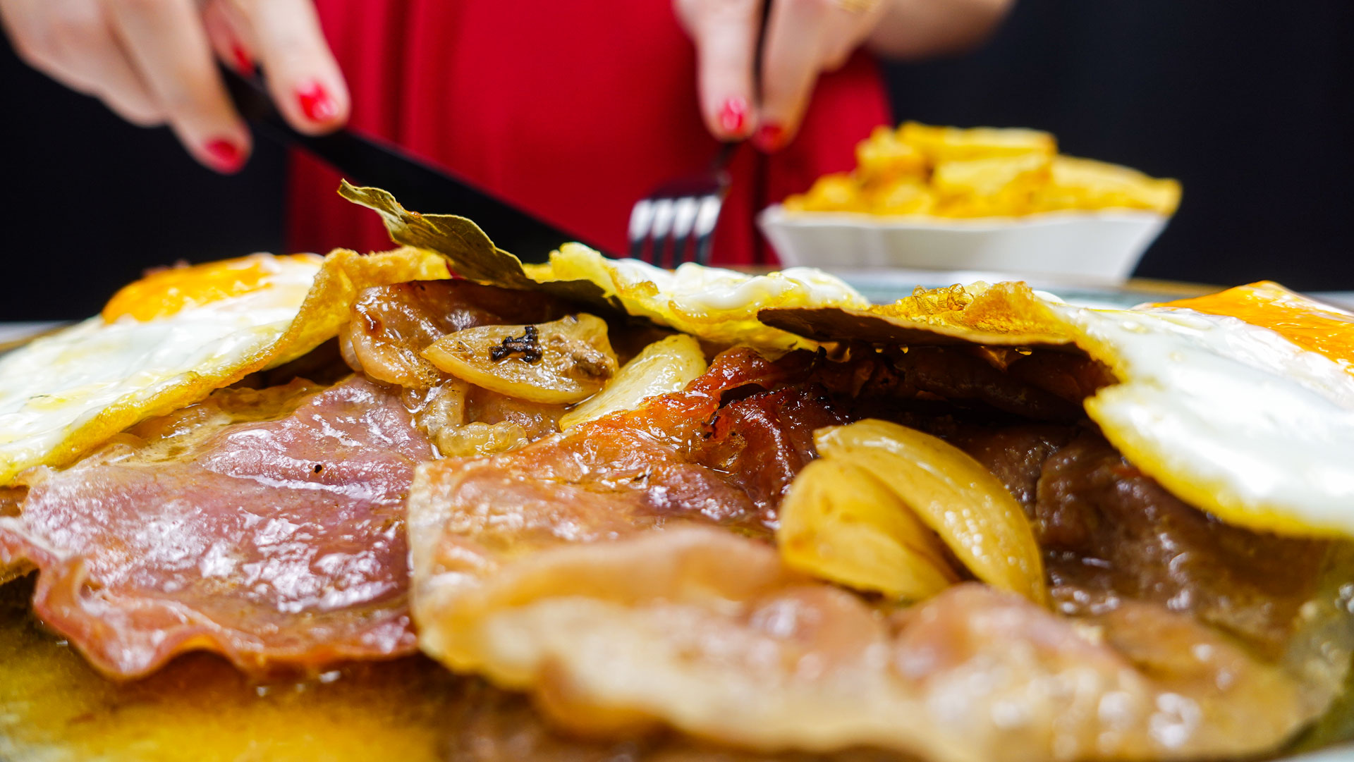 Helena is cutting steak with fried egg on a plate with fried potatoes in a bowl