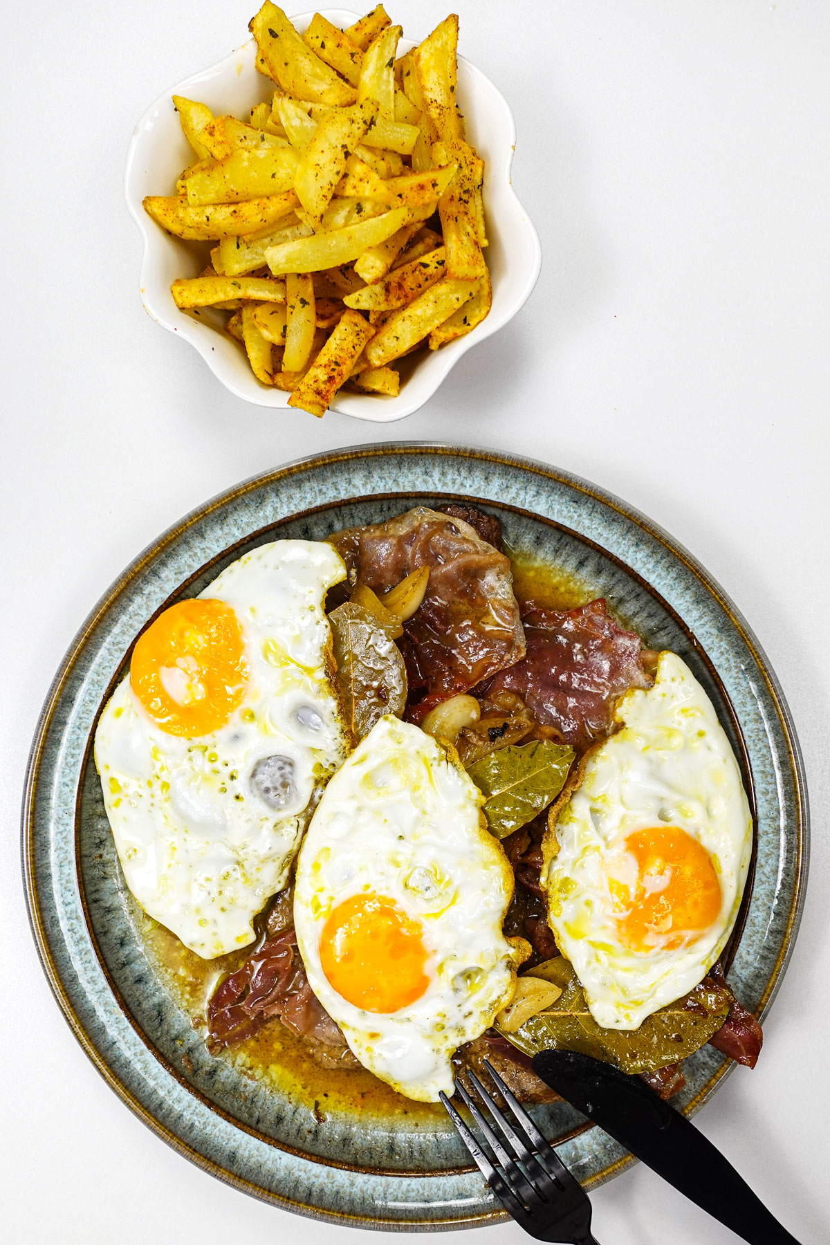 A plate wit 3 steaks with eggs on top and a small bowl of fried potatoes