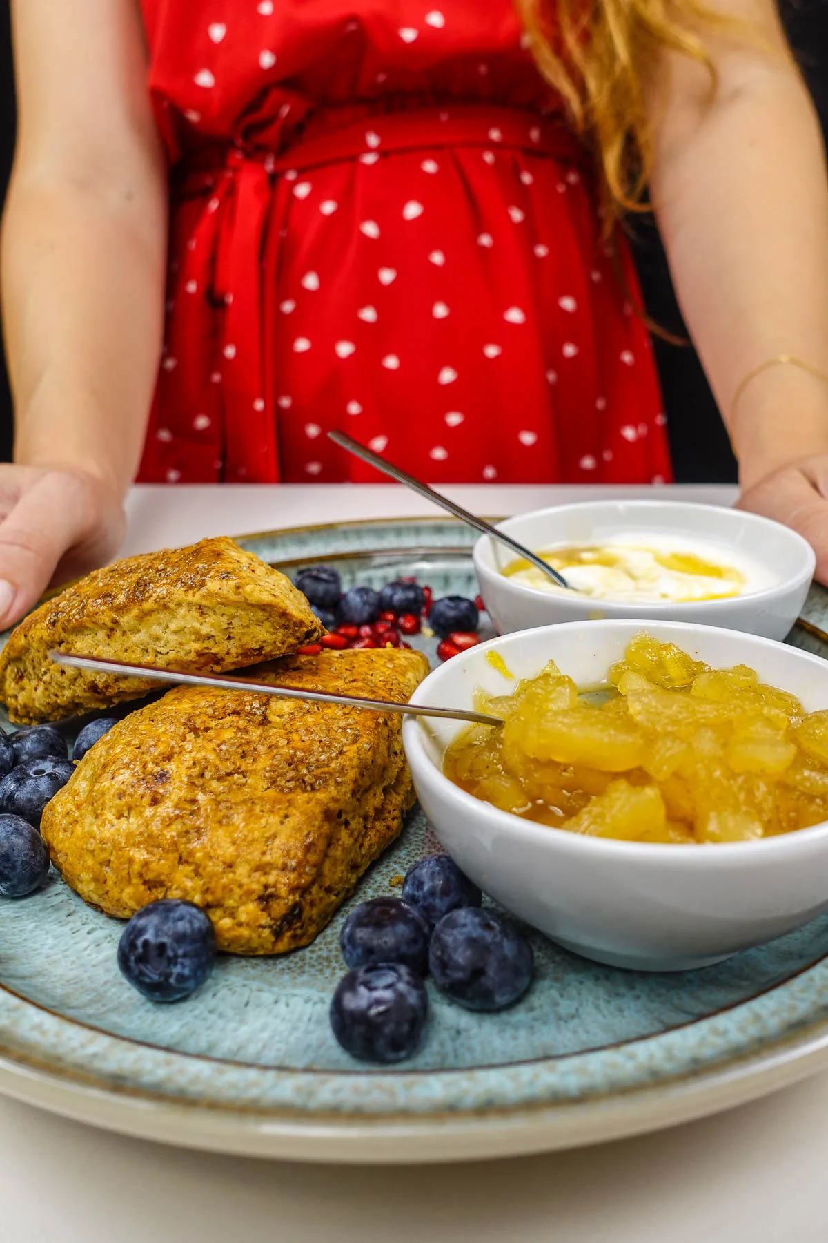 Helena is holding a plate with two cottage cheese scones, pear jam, some fresh blueberries and greek yogurt with honey.