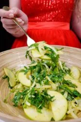 Helena is holding a fork full of juicy fennel apple salad with arugula in red dress