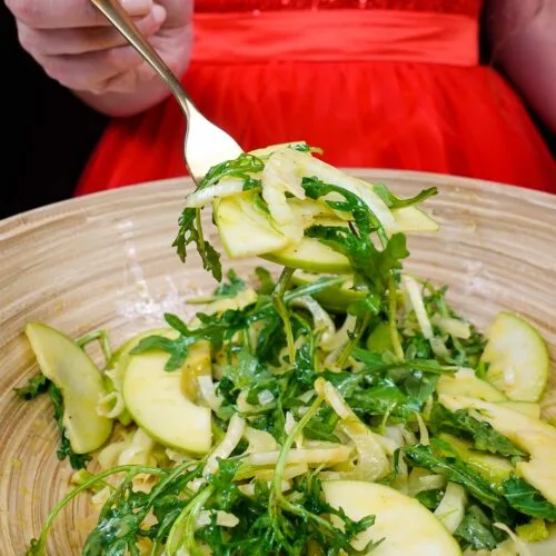 Helena is holding a fork full of juicy fennel apple salad with arugula in red dress