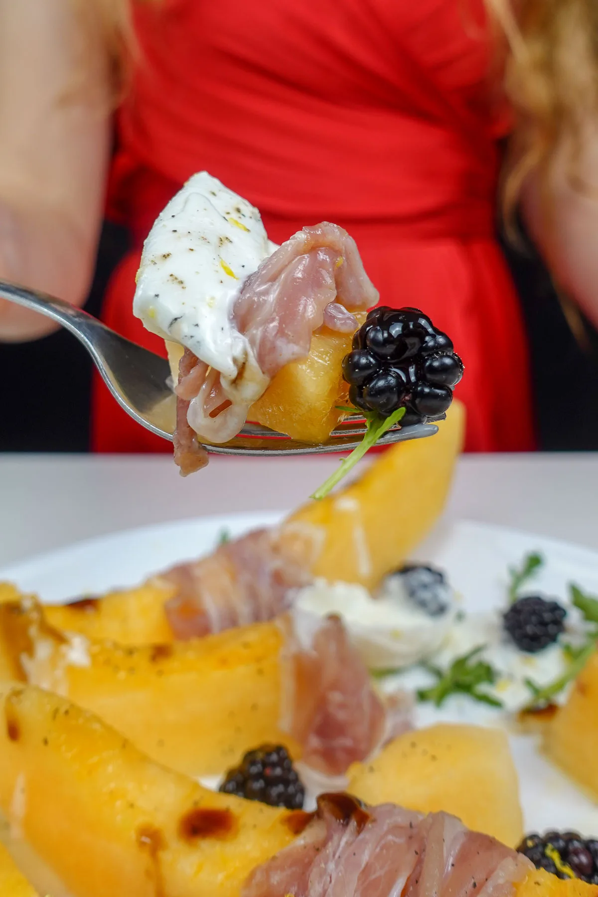 Helena is holding a fork full with melon prosciutto burrata and blackberries.