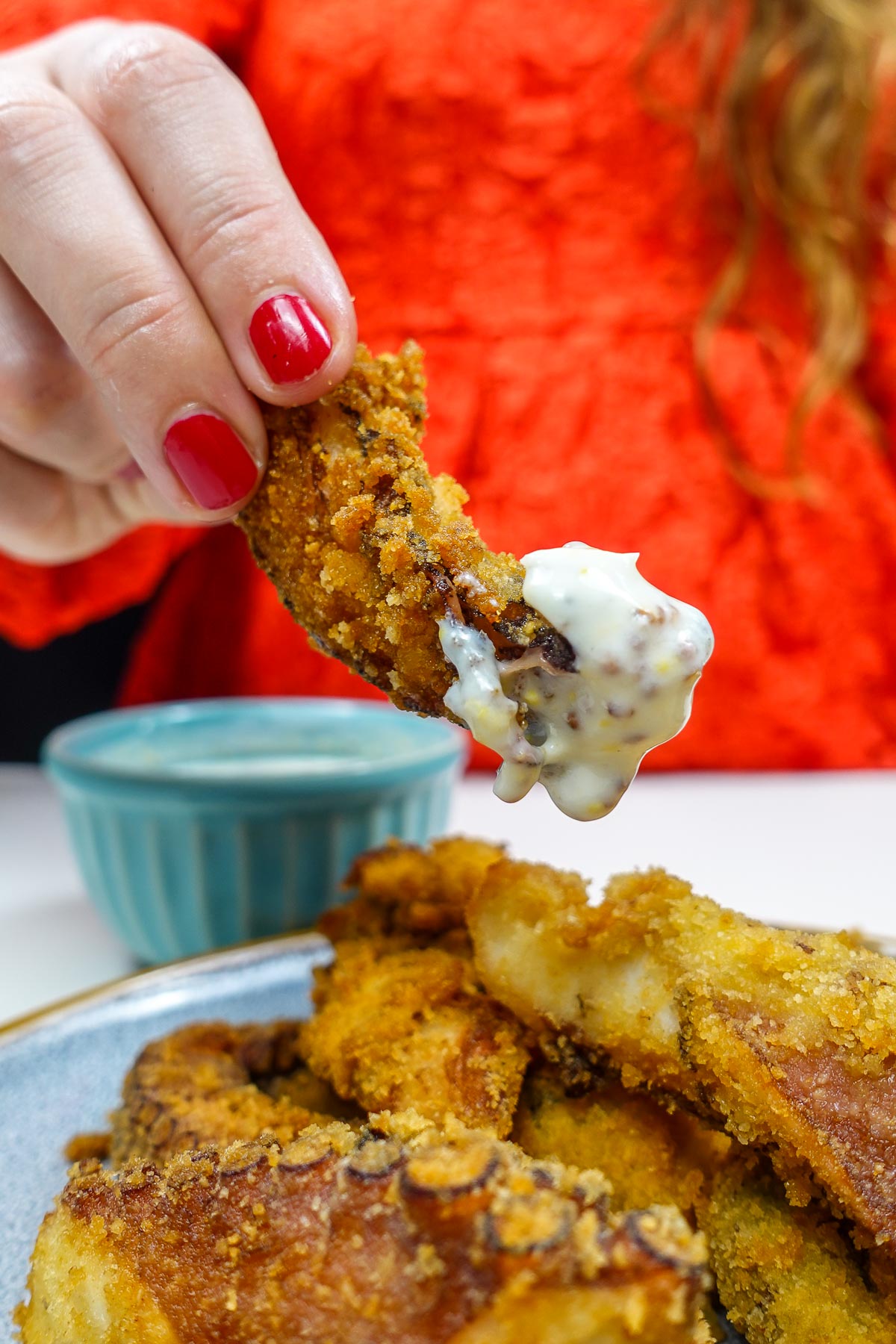 Helena is holding one piece of Portuguese fried octopus dipped in creamy sauce with plate of octopus.
