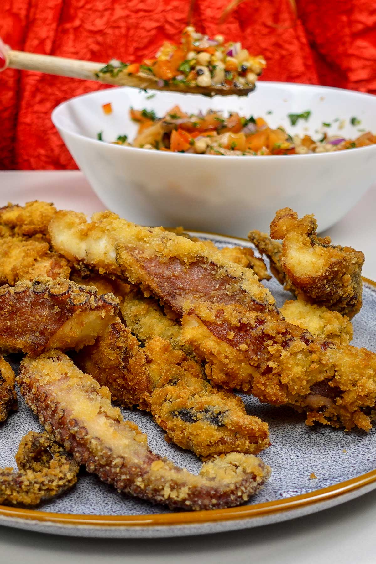 Helena is standing behind the plates with Portuguese Fried Octopus and holding a spoon bowl of vegetable salad.