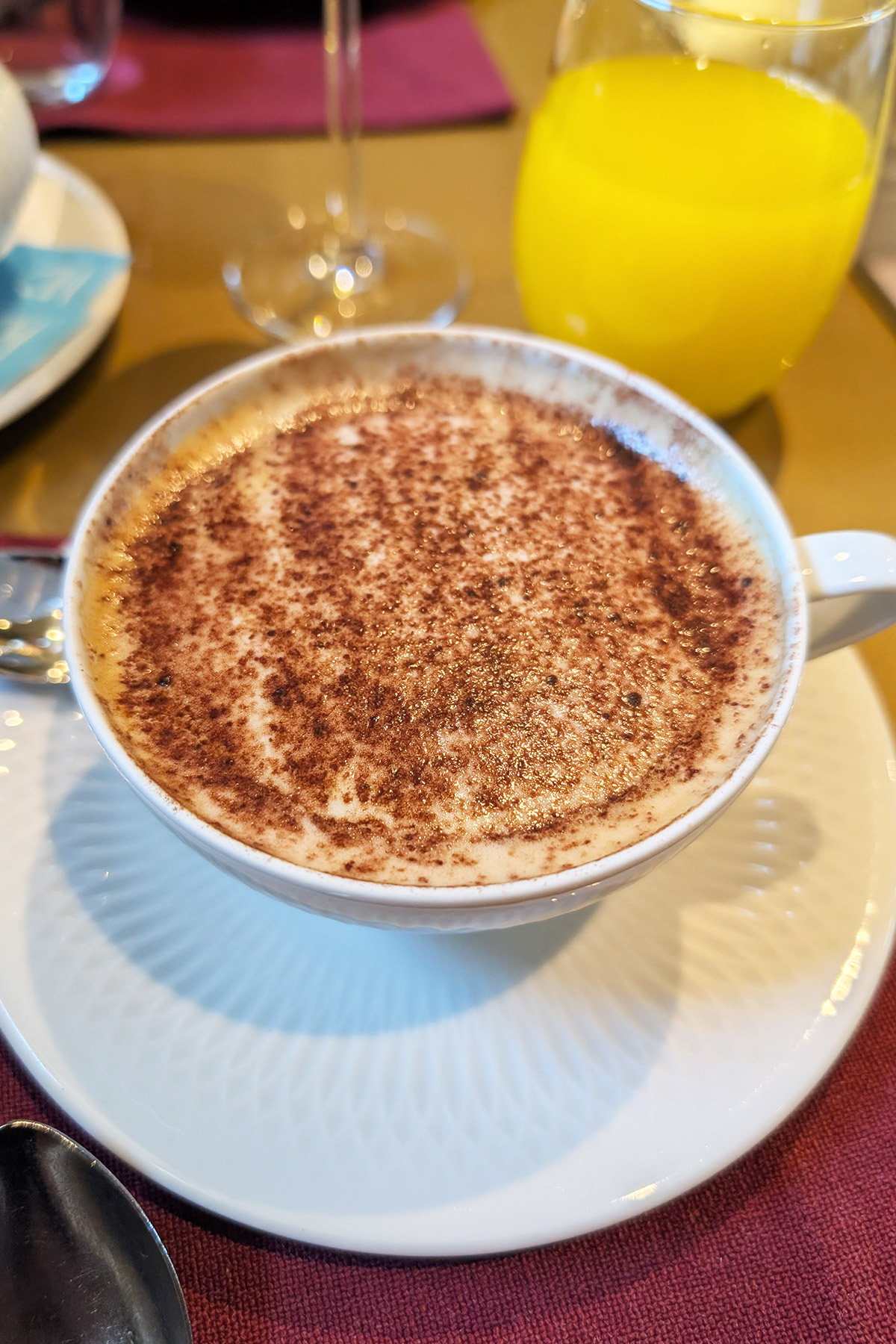 Cup of frothy cappuccino with cocoa powder on top and glass of orange juice.