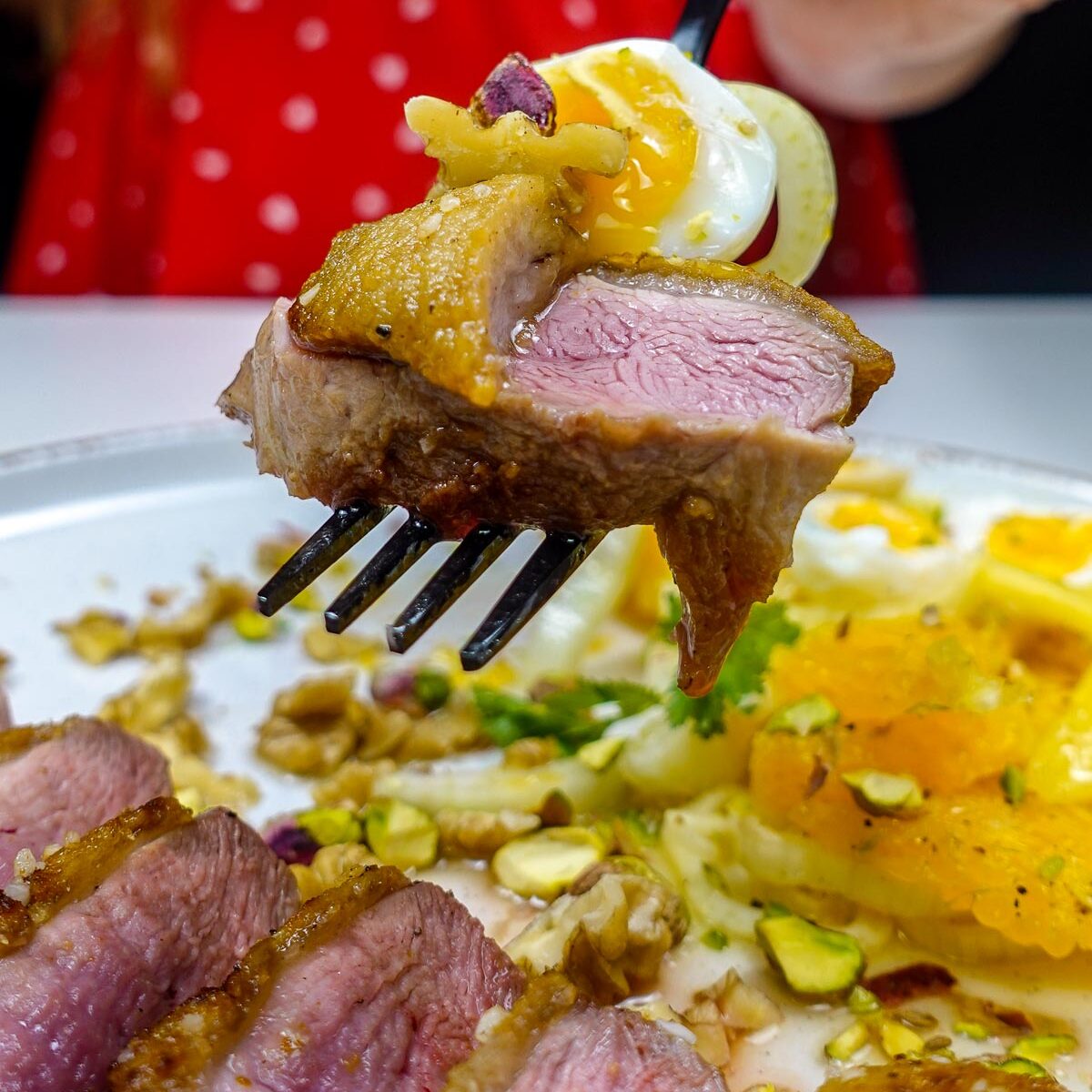 Helena is holding a black fork with crispy duck breast with soft boiled egg and fennel orange salad.
