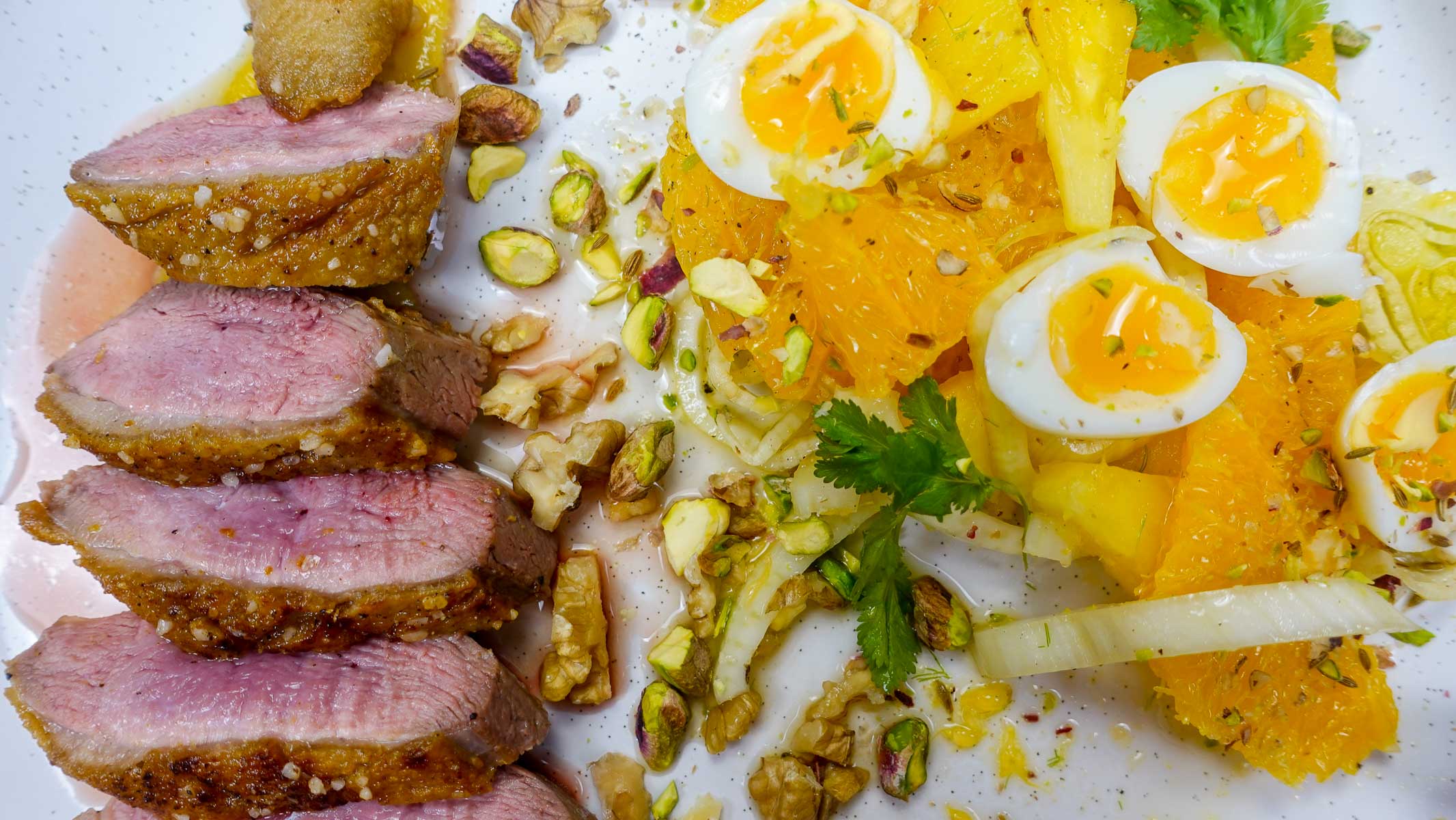 Sliced crispy duck breast served with orange, pineapple, soft boiled eggs, and nuts on a plate.