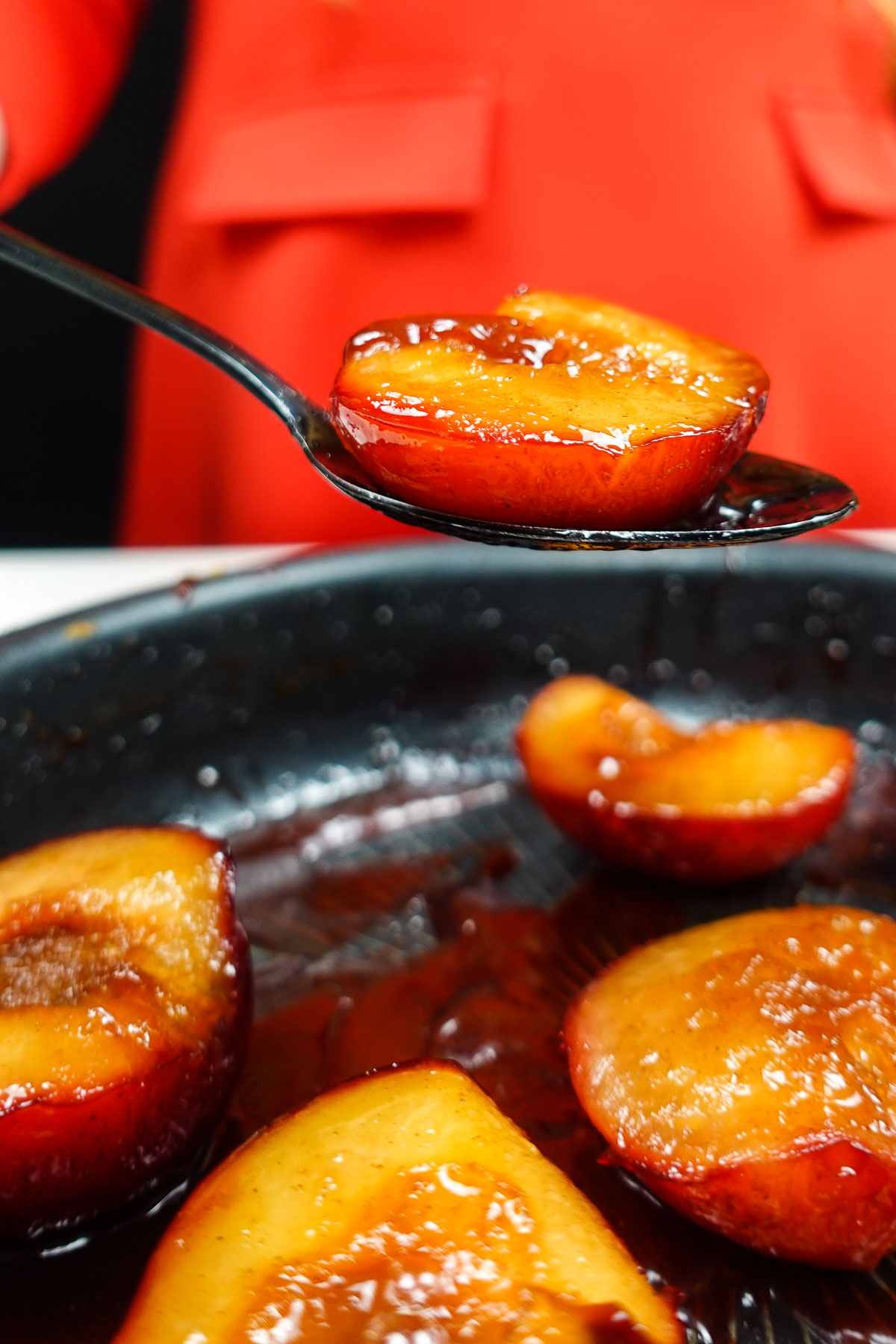 Caramelized plum half on spoon with plums in pan below.